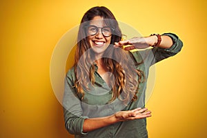 Young beautiful woman wearing green shirt and glasses over yelllow isolated background gesturing with hands showing big and large