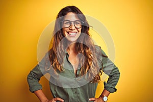 Young beautiful woman wearing green shirt and glasses over yelllow isolated background with a happy and cool smile on face