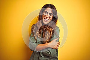 Young beautiful woman wearing green shirt and glasses over yelllow isolated background happy face smiling with crossed arms