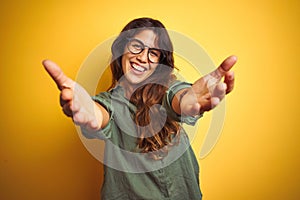 Young beautiful woman wearing green shirt and glasses over yelllow isolated background looking at the camera smiling with open