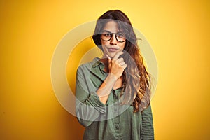 Young beautiful woman wearing green shirt and glasses over yelllow isolated background looking confident at the camera with smile