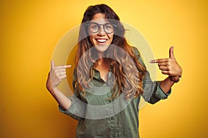 Young beautiful woman wearing green shirt and glasses over yelllow isolated background looking confident with smile on face,