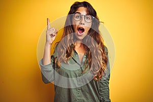 Young beautiful woman wearing green shirt and glasses over yelllow isolated background pointing finger up with successful idea