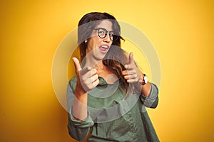 Young beautiful woman wearing green shirt and glasses over yelllow isolated background pointing fingers to camera with happy and