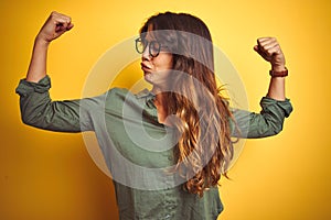 Young beautiful woman wearing green shirt and glasses over yelllow isolated background showing arms muscles smiling proud