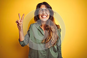 Young beautiful woman wearing green shirt and glasses over yelllow isolated background showing and pointing up with fingers number