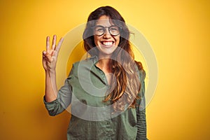 Young beautiful woman wearing green shirt and glasses over yelllow isolated background showing and pointing up with fingers number