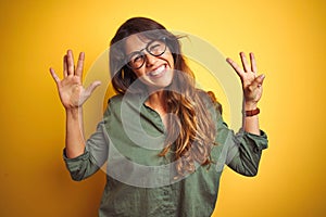 Young beautiful woman wearing green shirt and glasses over yelllow isolated background showing and pointing up with fingers number