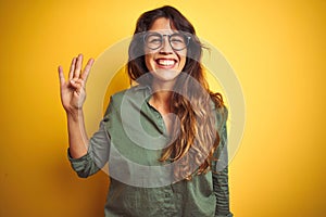 Young beautiful woman wearing green shirt and glasses over yelllow isolated background showing and pointing up with fingers number