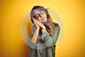 Young beautiful woman wearing green shirt and glasses over yelllow isolated background sleeping tired dreaming and posing with