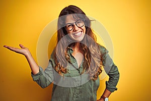 Young beautiful woman wearing green shirt and glasses over yelllow isolated background smiling cheerful presenting and pointing