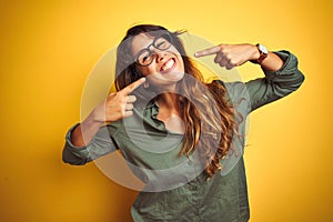 Young beautiful woman wearing green shirt and glasses over yelllow isolated background smiling cheerful showing and pointing with