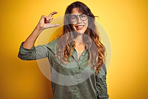Young beautiful woman wearing green shirt and glasses over yelllow isolated background smiling and confident gesturing with hand