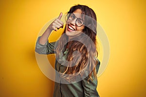 Young beautiful woman wearing green shirt and glasses over yelllow isolated background smiling doing phone gesture with hand and