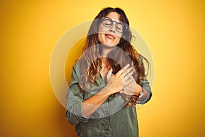 Young beautiful woman wearing green shirt and glasses over yelllow isolated background smiling with hands on chest with closed