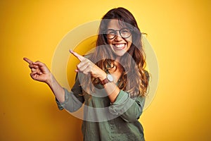 Young beautiful woman wearing green shirt and glasses over yelllow isolated background smiling and looking at the camera pointing