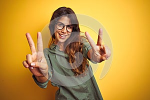 Young beautiful woman wearing green shirt and glasses over yelllow isolated background smiling looking to the camera showing