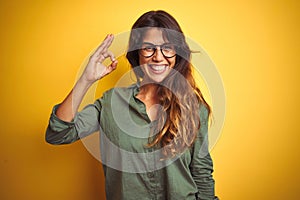 Young beautiful woman wearing green shirt and glasses over yelllow isolated background smiling positive doing ok sign with hand