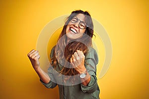 Young beautiful woman wearing green shirt and glasses over yelllow isolated background very happy and excited doing winner gesture