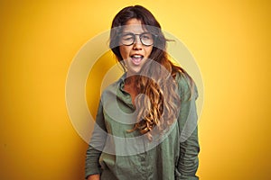 Young beautiful woman wearing green shirt and glasses over yelllow isolated background winking looking at the camera with sexy
