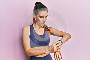 Young beautiful woman wearing gym clothes, using earphones and smart watch in shock face, looking skeptical and sarcastic,