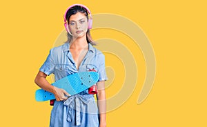 Young beautiful woman wearing headphones and holding skate thinking attitude and sober expression looking self confident