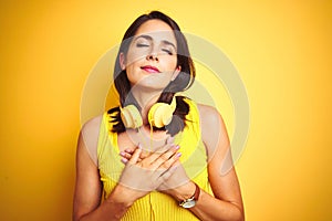 Young beautiful woman wearing headphones over yellow isolated background smiling with hands on chest with closed eyes and grateful