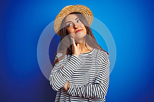 Young beautiful woman wearing navy striped t-shirt and hat over isolated blue background with hand on chin thinking about