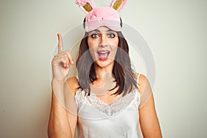 Young beautiful woman wearing pajama and mask standing over white isolated background pointing finger up with successful idea