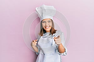 Young beautiful woman wearing professional cook uniform and hat pointing fingers to camera with happy and funny face