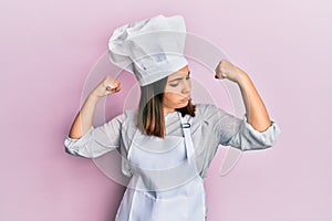 Young beautiful woman wearing professional cook uniform and hat showing arms muscles smiling proud