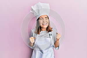 Young beautiful woman wearing professional cook uniform and hat very happy and excited doing winner gesture with arms raised,