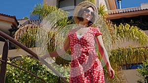 Young beautiful woman wearing red dress, hat and sunglasses standing on the bridge in the hotel garden with palm trees