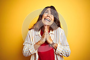 Young beautiful woman wearing red t-shirt and stripes shirt over yellow isolated background begging and praying with hands