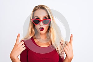 Young beautiful woman wearing red t-shirt and sunglasses over isolated white background amazed and surprised looking up and