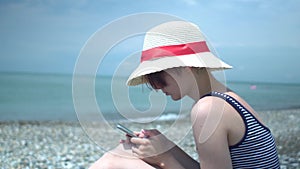 Young beautiful woman wearing retro hat and swimsuit with blue and white stripes in lonely summer beach using smartphone