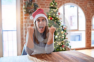 Young beautiful woman wearing santa claus hat at the table at home around christmas decoration celebrating surprised and amazed