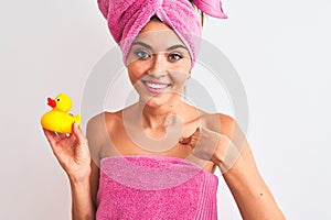 Young beautiful woman wearing shower towel holding duck over isolated white background with surprise face pointing finger to