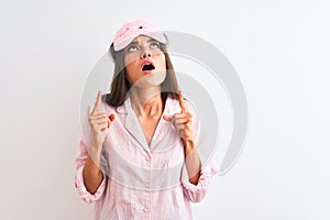 Young beautiful woman wearing sleep mask and pajama over isolated white background amazed and surprised looking up and pointing