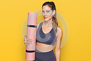 Young beautiful woman wearing sportswear holding yoga mat thinking attitude and sober expression looking self confident