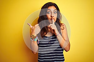 Young beautiful woman wearing striped t-shirt standing over isolated yellow background asking to be quiet with finger on lips