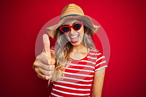 Young beautiful woman wearing sunglasses and summer hat over red isolated background doing happy thumbs up gesture with hand