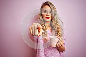 Young beautiful woman wearing sweater drinking a cup of coffee over pink isolated background pointing with finger to the camera