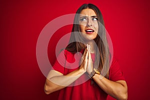 Young beautiful woman wearing t-shirt standing over isolated red background begging and praying with hands together with hope