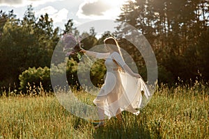 Young beautiful woman, wearing white dress, holding flowers and dancing on the meadow. Girl joying nature and freedom