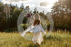 Young beautiful woman, wearing white dress, holding flowers and dancing on the meadow. Girl joying nature and freedom