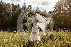 Young beautiful woman, wearing white dress, holding flowers and dancing on the meadow. Girl joying nature and freedom