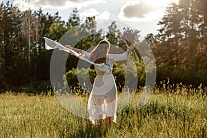 Young beautiful woman, wearing white dress, holding flowers and dancing on the meadow. Girl joying nature and freedom