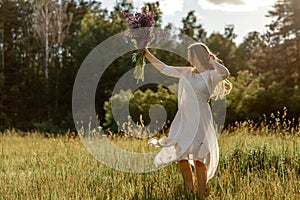 Young beautiful woman, wearing white dress, holding flowers and dancing on the meadow. Girl joying nature and freedom