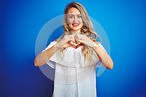 Young beautiful woman wearing white dress standing over blue isolated background smiling in love showing heart symbol and shape
